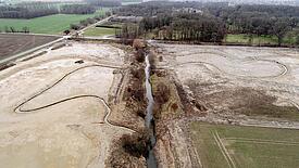 Luftaufnahme einer sandigen Fläche durch die ein gewundener Bach fließt, In der Mitte ein Graben, im Hintergrund Bäume ohne Laub