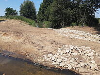 Im Vordergrund steht Wasser, dahinter ein Damm aus Erde und SSteinen, im Hintergrund fließt ein Bach zwischen grünen Bäumen.